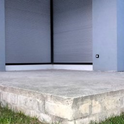 MATEUSZ KALISZUK TechRol - Szare, poziome lamele osłonowe na oknach nowoczesnego budynku z betonowym podwyższeniem i trawnikiem. Minimalistyczny design, stonowana kolorystyka.