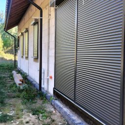 MATEUSZ KALISZUK TechRol - Szare rolety na oknach surowego budynku z betonu komórkowego, widok z zewnątrz. Paleta drewniana na pierwszym planie, trawa i krzewy w tle.