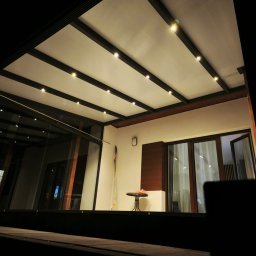 MATEUSZ KALISZUK TechRol - Nowoczesna, oświetlona pergola z metalową konstrukcją i szklanymi ścianami, widok z dołu, nocne oświetlenie punktowe, minimalistyczny design.