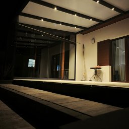 MATEUSZ KALISZUK TechRol - Nowoczesna, oświetlona pergola z przeszklonymi ścianami, widoczna z poziomu schodów. Na ścianie akcent dekoracyjny i stolik. Ujęcie nocne.