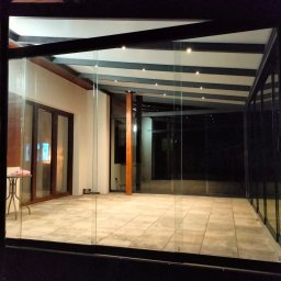 MATEUSZ KALISZUK TechRol - Szklana zabudowa tarasu z drewnianą konstrukcją dachu i oświetleniem punktowym, widok z zewnątrz w nocy. Widoczne płytki podłogowe i częściowo meble.