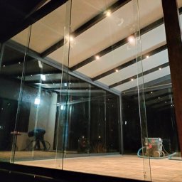 MATEUSZ KALISZUK TechRol - Szklana zabudowa tarasu z metalową konstrukcją, oświetlona punktowo. Widoczne odbicia otoczenia i osoba wewnątrz. Podłoga wyłożona płytkami.