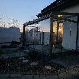 MATEUSZ KALISZUK TechRol - Szklana zabudowa balkonu z metalową konstrukcją, widoczna z zewnątrz o zmierzchu. Na pierwszym planie kamienna ścieżka i fragment ogrodu, w tle biały bus.