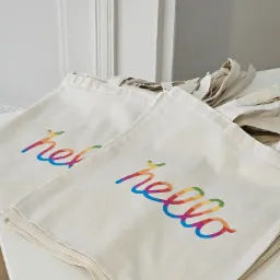 Stos beżowych toreb bawełnianych z nadrukowanym kolorowym napisem 'hello' na białym stole.