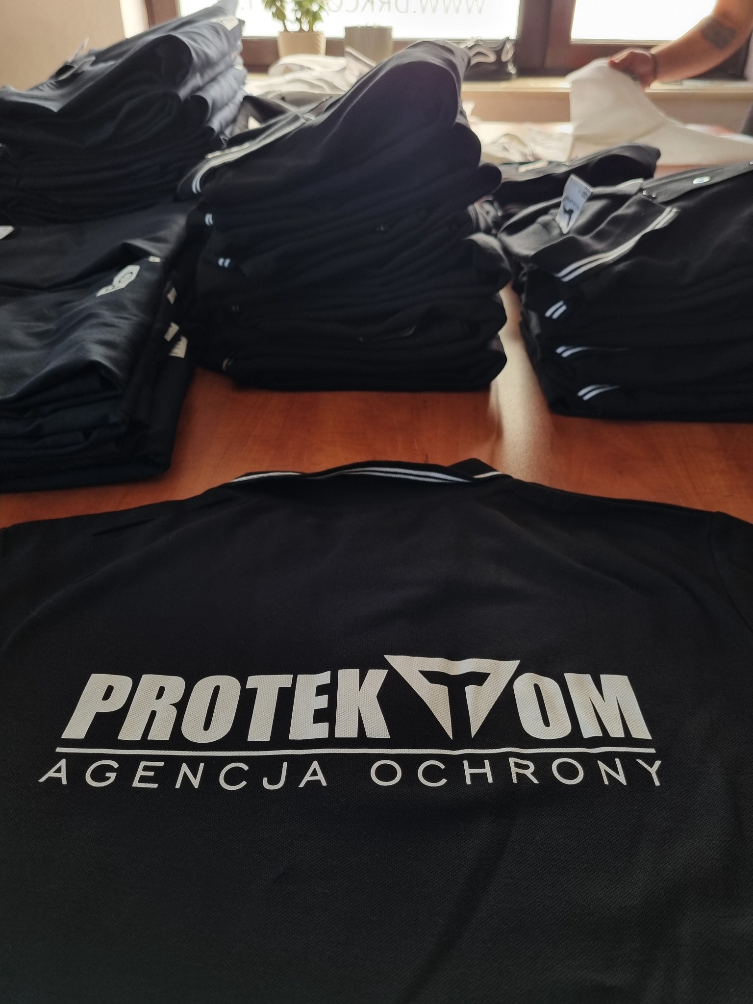 Stos czarnych koszulek polo z białym logo 'PROTEKTOM Agencja Ochrony' na stole, z kolejnymi koszulkami w tle.