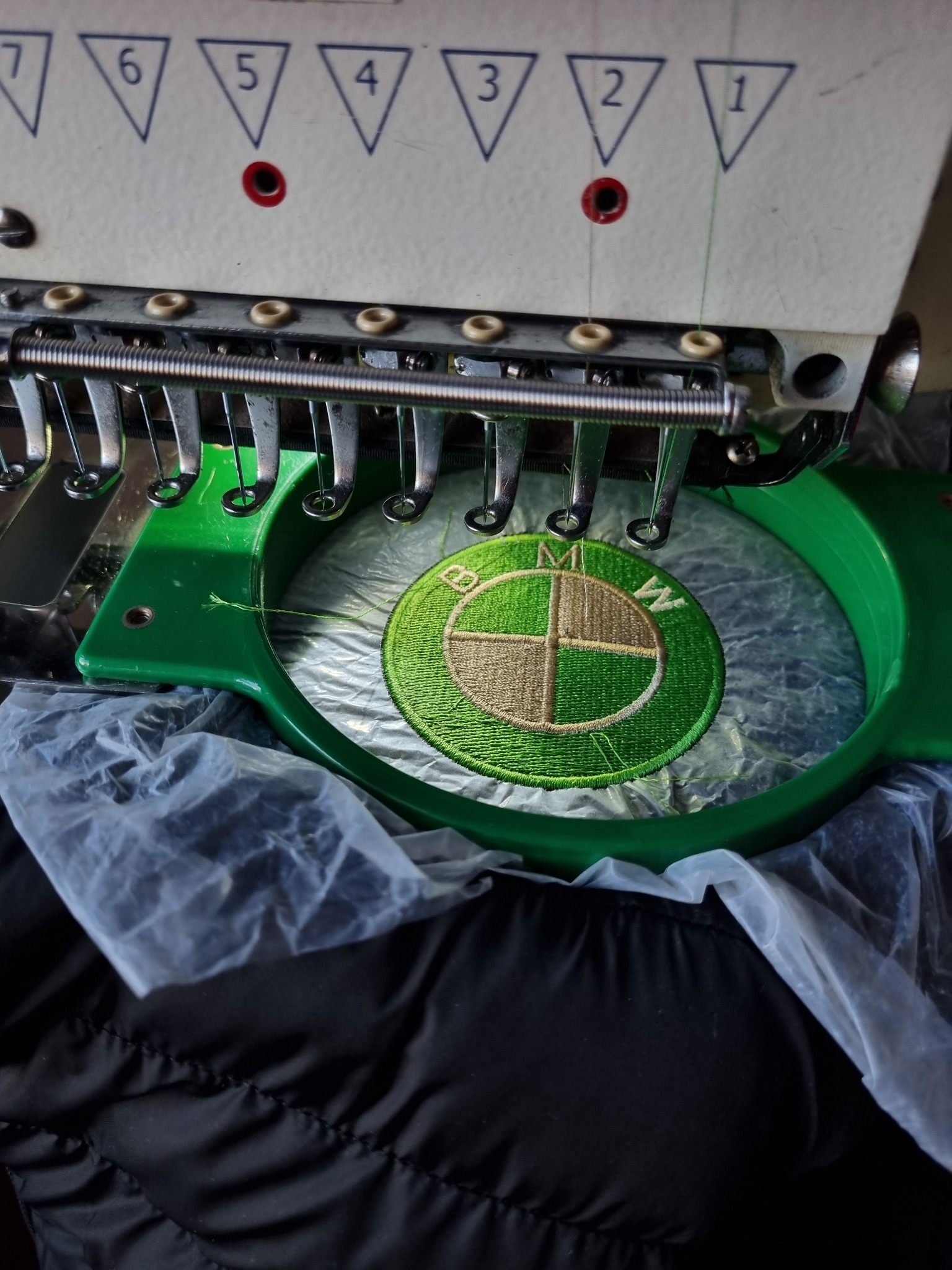 Proces haftowania logo motoryzacyjnego na materiale, widoczne igły maszyny i zielony tamborek, częściowo widoczna czarna kurtka