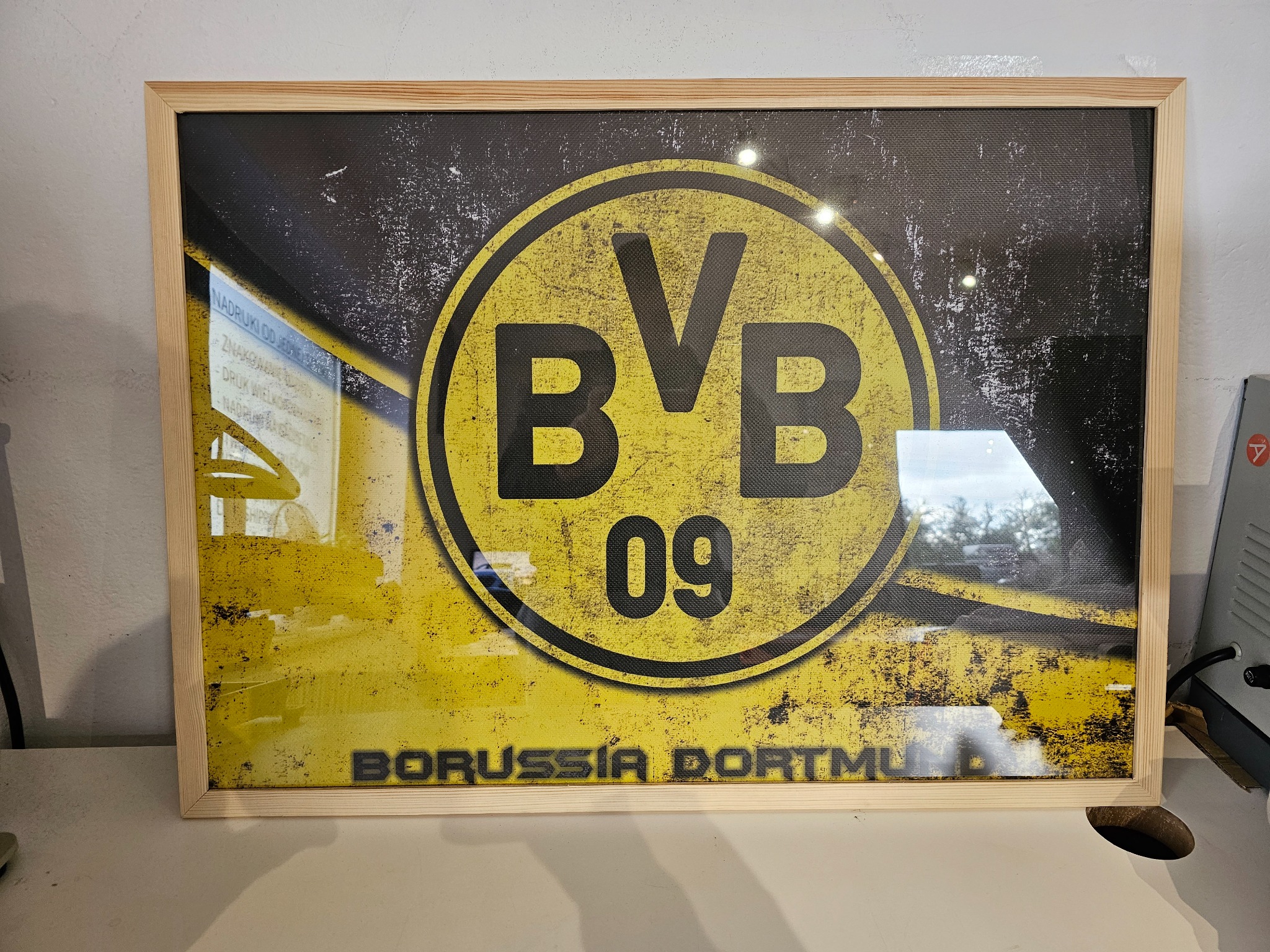 Oprawiony w drewnianą ramę plakat z logo klubu Borussia Dortmund (BVB 09), stylizowany na vintage, z widocznymi śladami użytkowania i blikami światła na szkle.