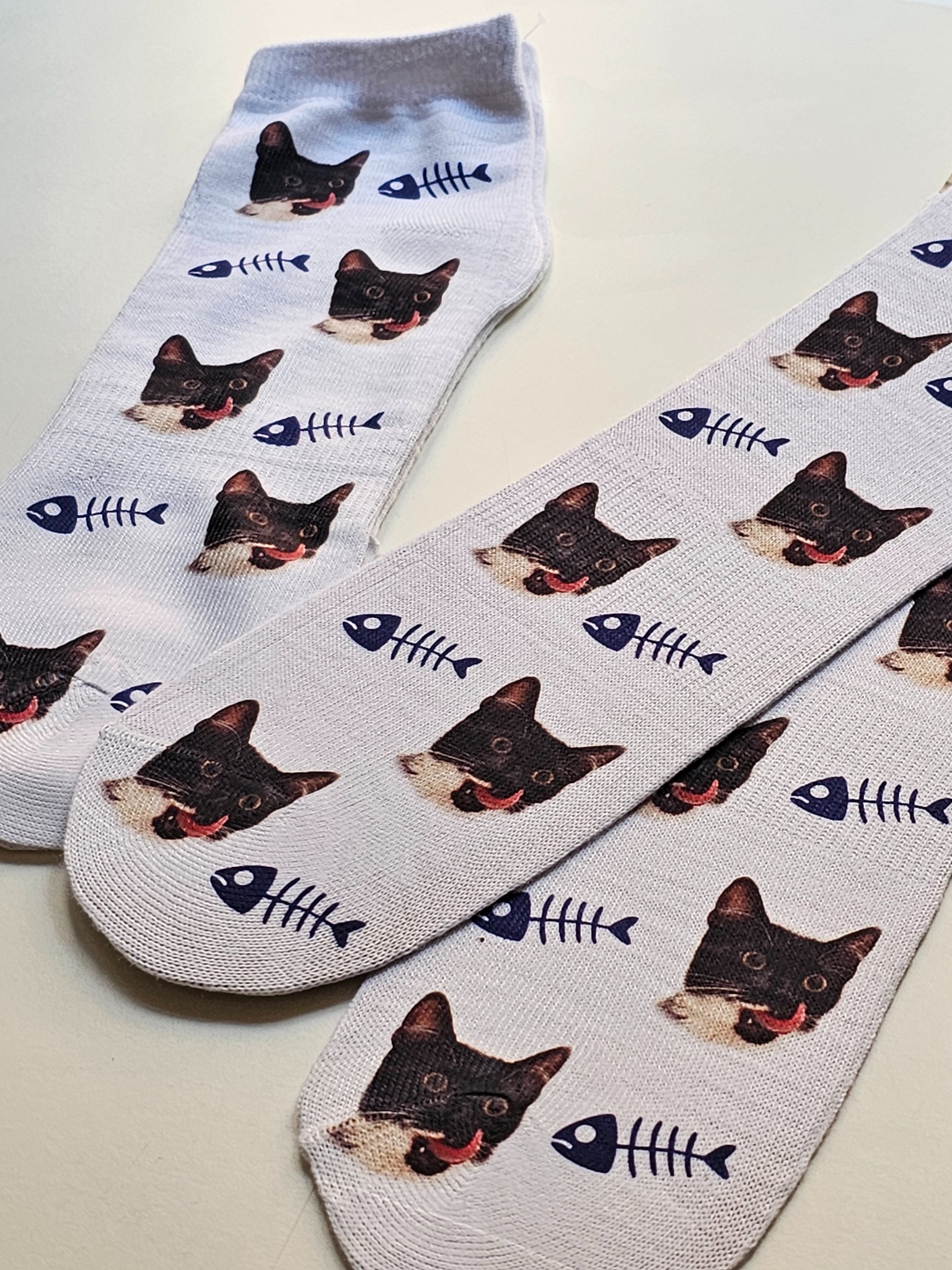 Dwie białe skarpety z powtarzającym się wzorem głowy czarno-białego kota z wyciągniętym językiem oraz granatowych szkieletów ryb.