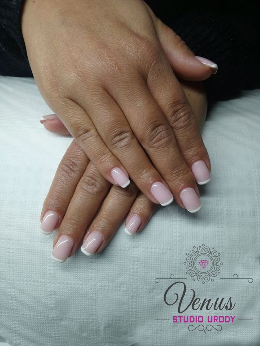 Dłonie z delikatnym, jasnoróżowym manicure french na krótkich paznokciach, logo salonu urody w prawym dolnym rogu.