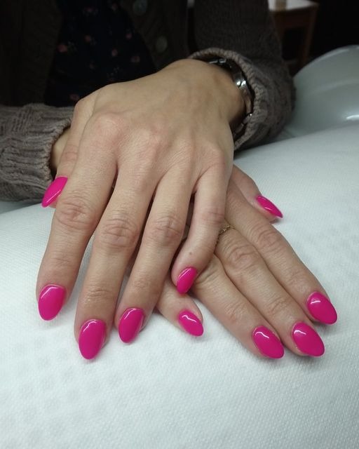 Dłonie z świeżo pomalowanymi, intensywnie różowymi paznokciami o migdałowym kształcie, delikatny pierścionek na jednym palcu.