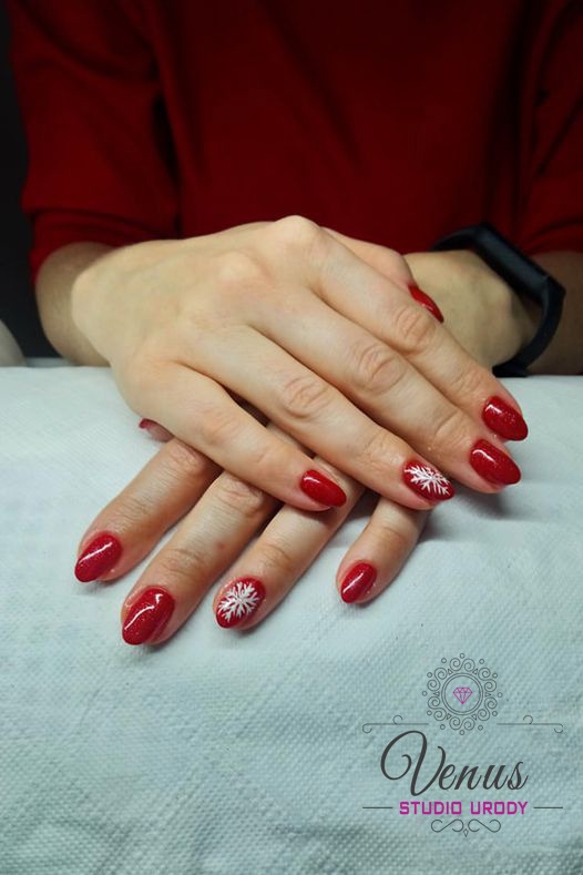 Dłonie kobiety z czerwonym, błyszczącym manicure hybrydowym i białymi wzorami śnieżynek na dwóch paznokciach, na tle czerwonej bluzki i logo salonu Venus.