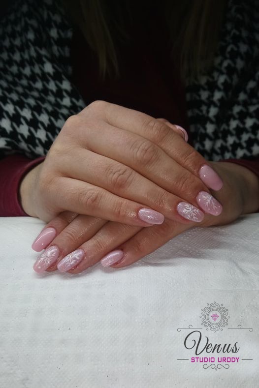 Dłonie kobiety z delikatnym, różowym manicure hybrydowym ozdobionym białymi wzorami śnieżynek na kilku paznokciach, ułożone na białym ręczniku z widocznym logo salonu urody.