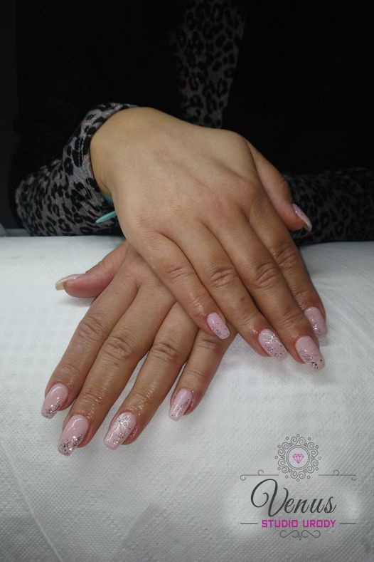 Dłonie kobiety z delikatnym, różowym manicure z brokatowymi akcentami i subtelnym wzorem kwiatowym na jednym paznokciu, ułożone na białym ręczniku z widocznym logo salonu urody.