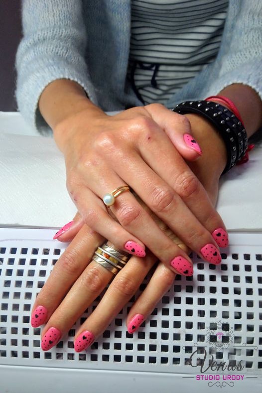 Ręce kobiety z różowym manicurem zdobionym czarnymi serduszkami, pierścionki na palcach, czarna bransoletka z ćwiekami, na tle białego podgrzewacza do rąk.
