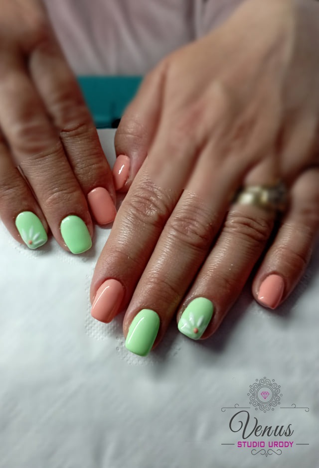 Dłonie z pomalowanymi paznokciami w odcieniach pastelowej zieleni i brzoskwini, na dwóch paznokciach zielonych namalowane białe kwiatki z pomarańczowymi środkami, na palcu serdecznym widoczny złoty...