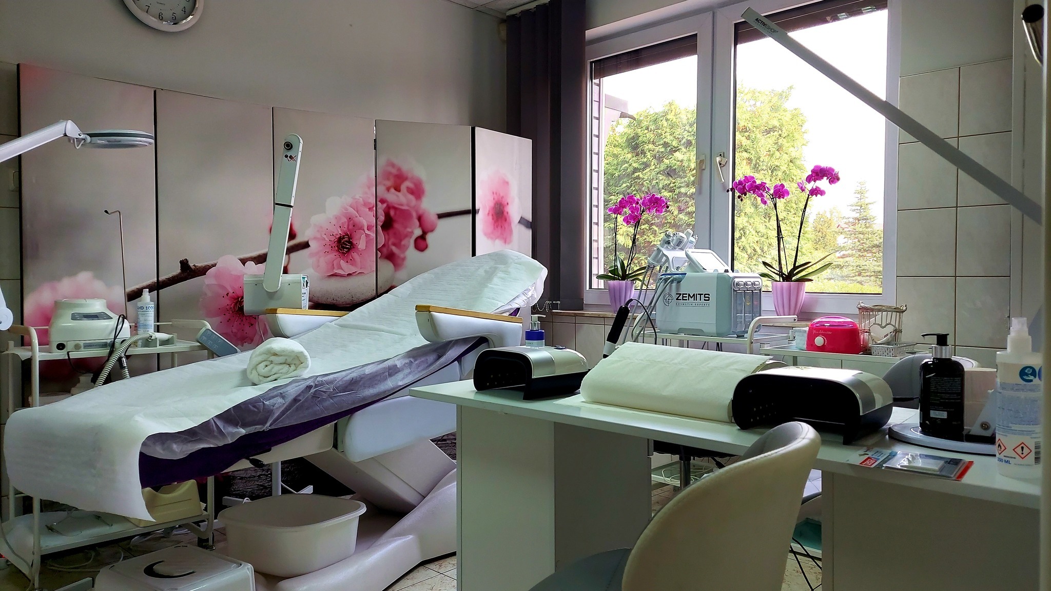 Jasne wnętrze gabinetu kosmetycznego z łóżkiem do zabiegów, lampą kosmetyczną i urządzeniami, w tle okno z widokiem na zieleń i storczyki w doniczkach.