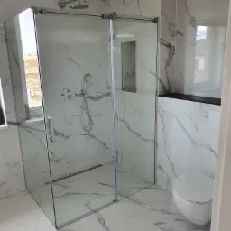 Nowoczesna kabina prysznicowa z szklanymi drzwiami przesuwnymi, wykończona płytkami imitującymi marmur, z widoczną armaturą i wnęką w ścianie, oraz toaleta w tle.