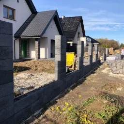 Wznoszenie ogrodzenia z betonowych bloczków modułowych na posesji we Wrocławiu, widoczne słupki i podmurówka w trakcie budowy, w tle dwa domy jednorodzinne i dwaj mężczyźni.