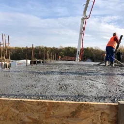 Wylewanie betonu na zbrojony fundament, widoczna pompa do betonu, pracownik w kamizelce odblaskowej rozprowadzający beton, szalunek z płyty OSB w pierwszym planie.