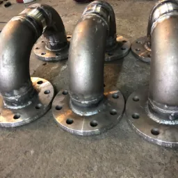 Trzy spawane kolanka stalowe z kołnierzami, widoczne ślady spawania i napisy na kołnierzach, leżące na szarej, betonowej powierzchni.