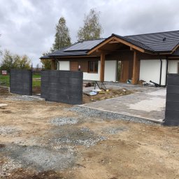 Odm.group Sp.z.o.o - Nowoczesna posesja z szarą, betonową bramą wjazdową, częściowo wykończonym podjazdem z kostki i domem z panelami słonecznymi na dachu w tle, widok na plac budowy.