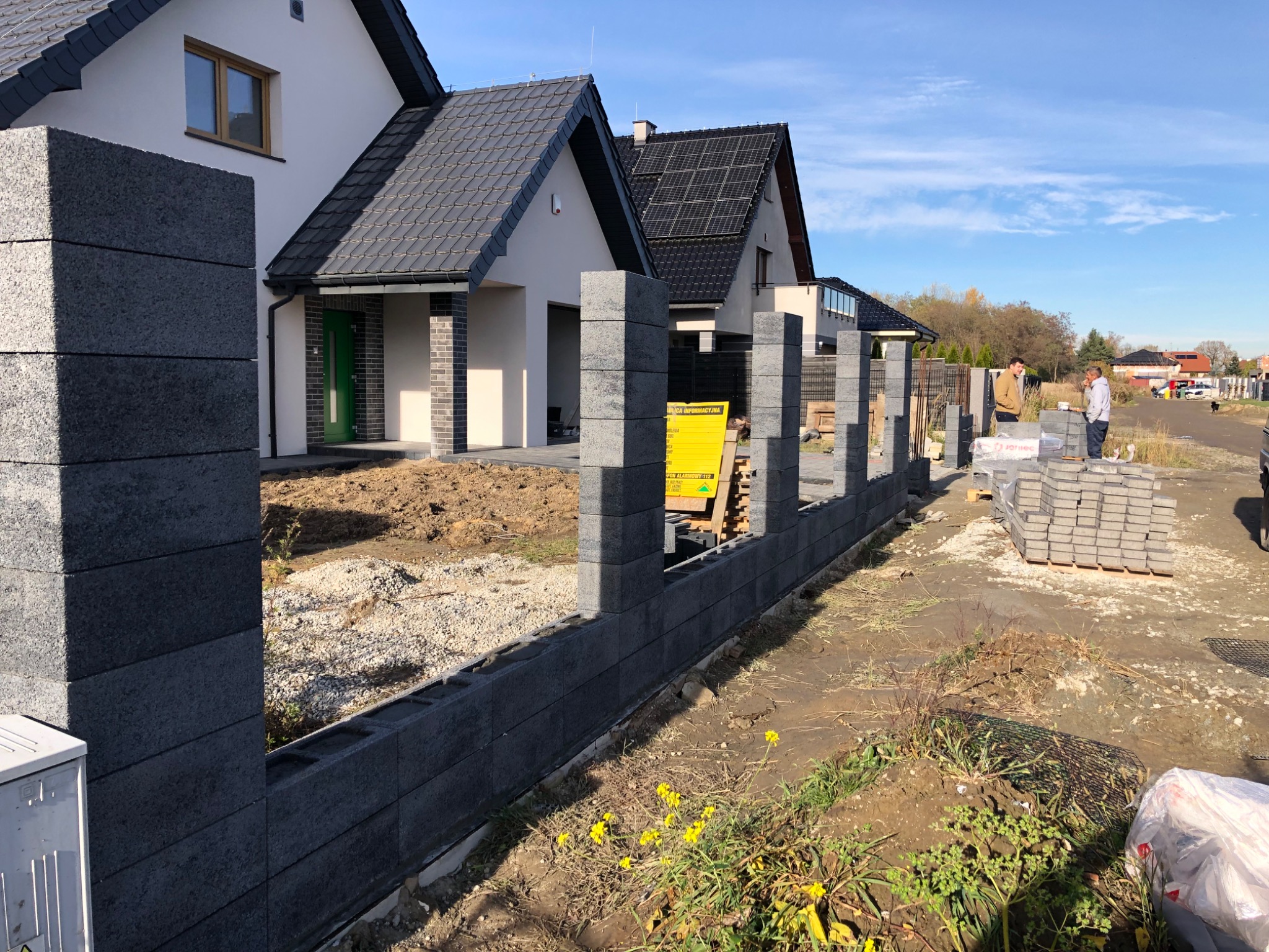 Wznoszenie ogrodzenia z betonowych bloczków modułowych na posesji we Wrocławiu, widoczne słupki i podmurówka w trakcie budowy, w tle dwa domy jednorodzinne i dwaj mężczyźni.