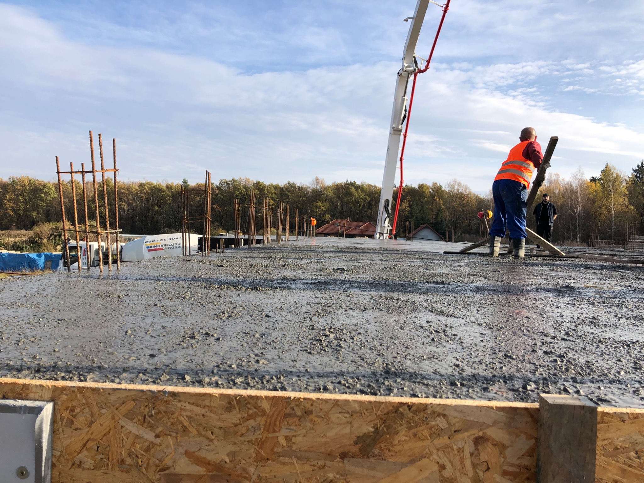 Wylewanie betonu na zbrojony fundament, widoczna pompa do betonu, pracownik w kamizelce odblaskowej rozprowadzający beton, szalunek z płyty OSB w pierwszym planie.