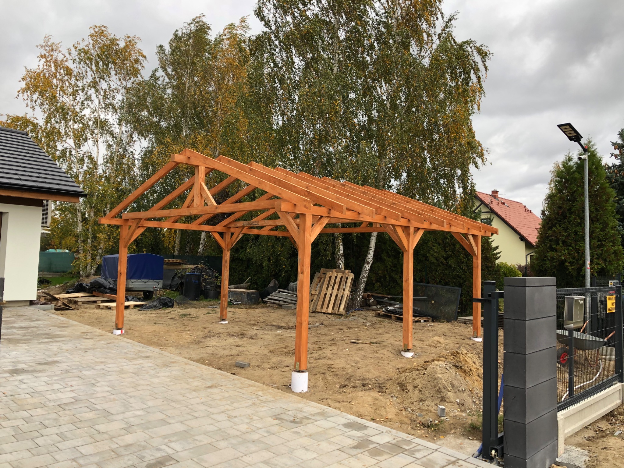 Drewniana konstrukcja zadaszenia w trakcie budowy, widoczne słupy i krokwie, w tle dom z ciemną dachówką i drzewa liściaste, obok kostka brukowa i częściowo widoczne ogrodzenie.