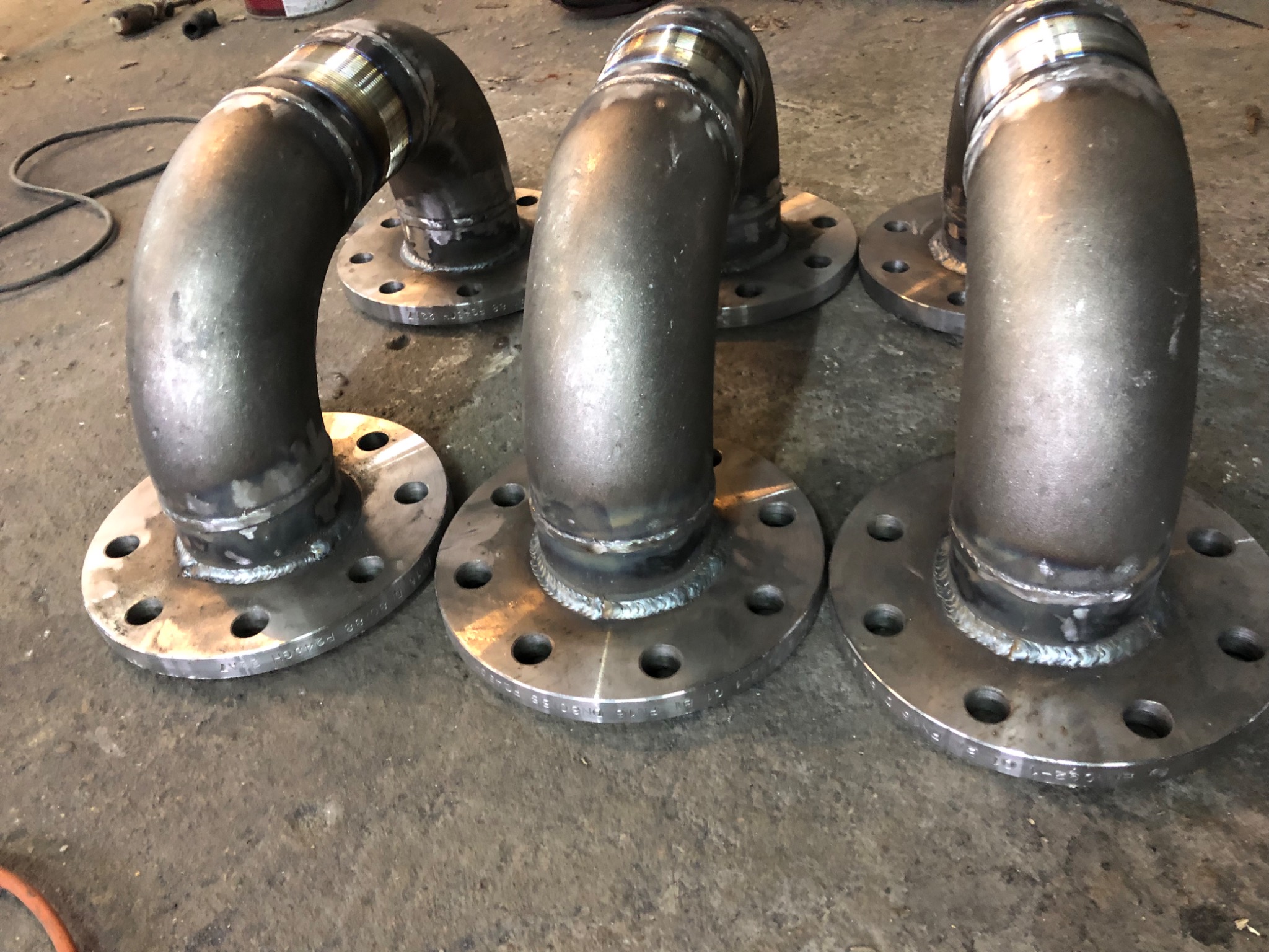 Trzy spawane kolanka stalowe z kołnierzami, widoczne ślady spawania i napisy na kołnierzach, leżące na szarej, betonowej powierzchni.