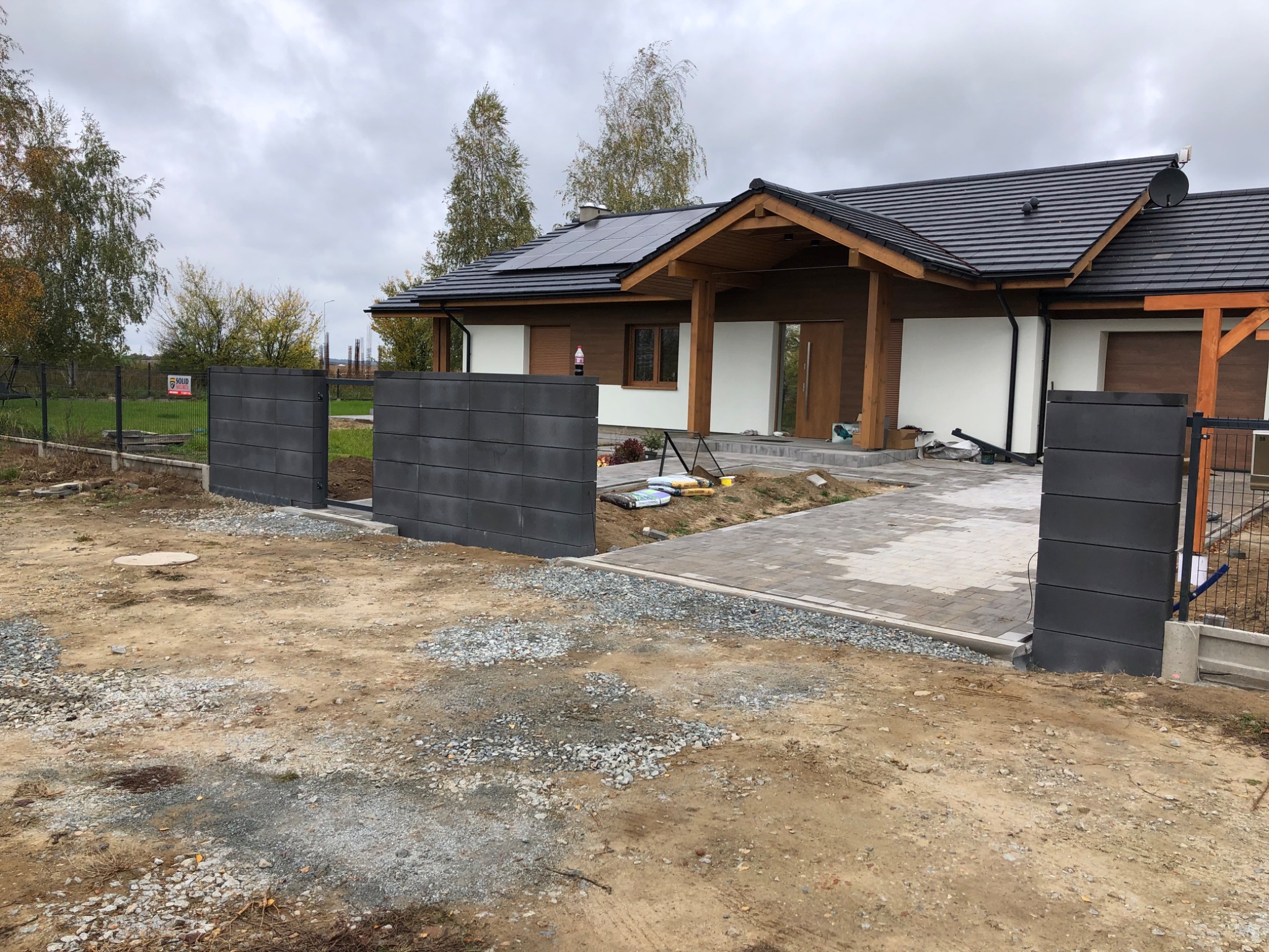 Nowoczesna posesja z szarą, betonową bramą wjazdową, częściowo wykończonym podjazdem z kostki i domem z panelami słonecznymi na dachu w tle, widok na plac budowy.