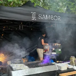 Czarny namiot z logo 'Sambor' podczas przygotowywania jedzenia na grillu, widoczny dym i osoba pracująca wewnątrz, oświetlenie dekoracyjne w tle.
