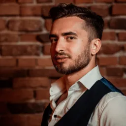 Portret młodego mężczyzny w białej koszuli i granatowej kamizelce na tle ceglanej ściany, z delikatnym uśmiechem i spojrzeniem skierowanym w bok.