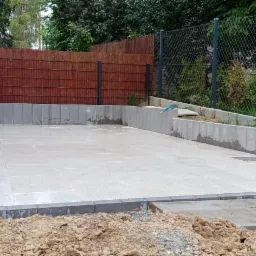 Wyłożony jasnymi płytami plac z obrzeżami z szarej kostki, widoczny fragment ziemi z przodu, w tle ogrodzenie z siatki i drewniana osłona, mokra powierzchnia po deszczu.