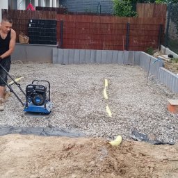 Scann Piotr Froń - Pracownik ubija zagęszczarką wibracyjną podłoże z kamieni pod przyszłą nawierzchnię, widoczne krawężniki i obrzeża, przygotowanie terenu przed domem.