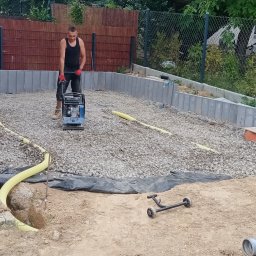 Scann Piotr Froń - Prace ziemne: mężczyzna w roboczych butach i czerwonych rękawicach zagęszcza podłoże z kamieni za pomocą zagęszczarki wibracyjnej, widoczne rury i krawężniki.