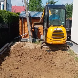 Żółta mini koparka JCB na placu budowy, obok czarnego ogrodzenia i rur kanalizacyjnych, na tle białej ściany budynku i drewnianej szopy.