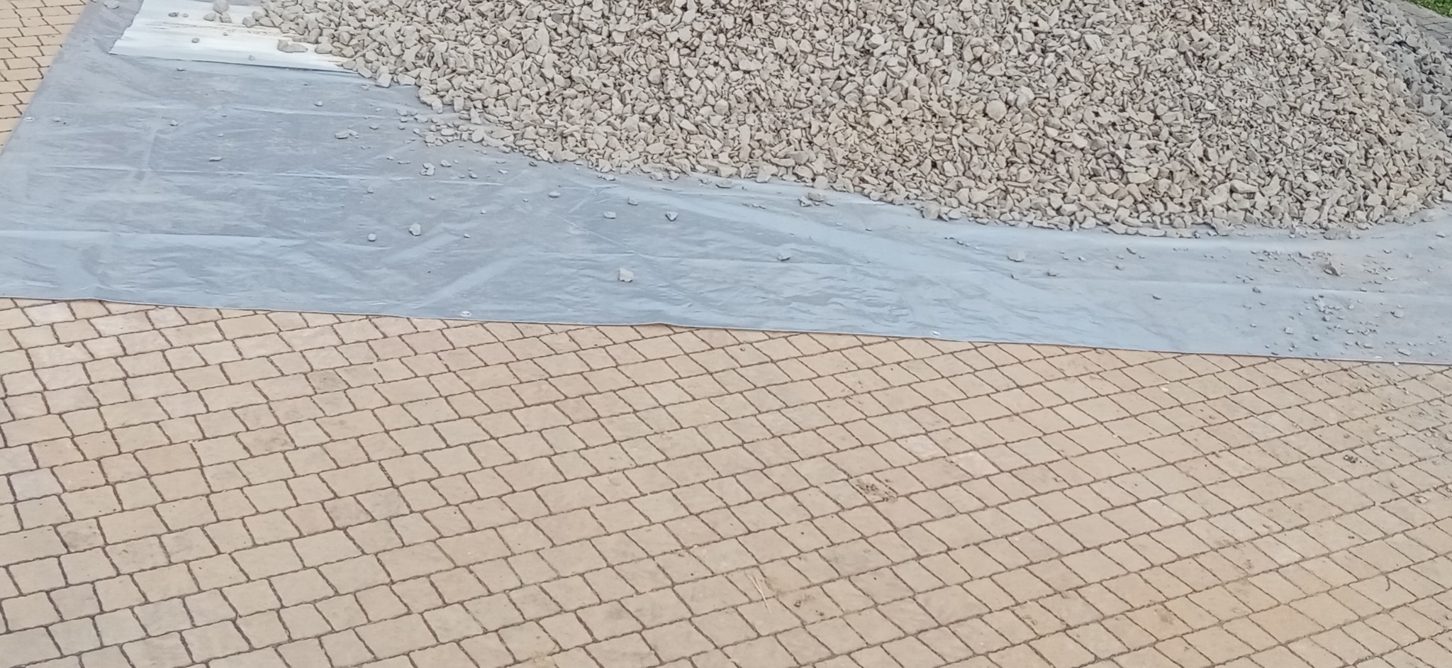 Stos białego kruszywa na szarej folii, częściowo rozsypany na świeżo ułożonej kostce brukowej w odcieniach beżu.