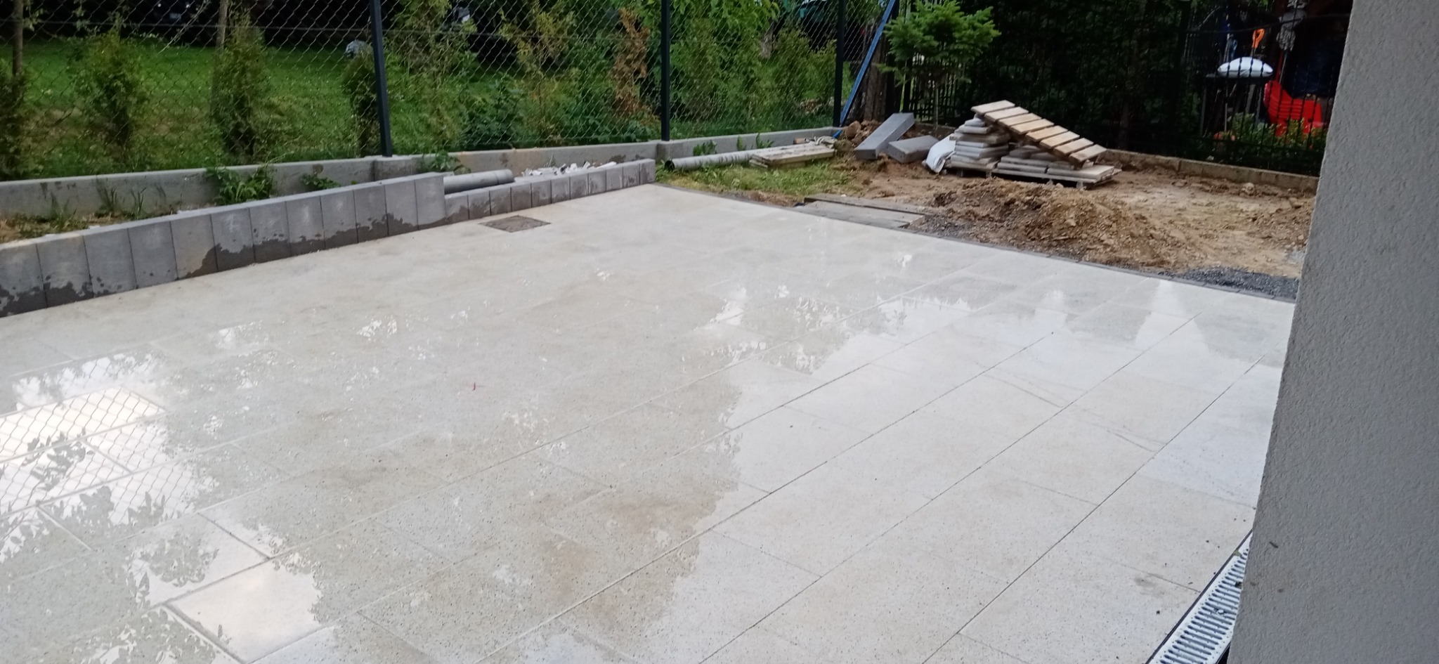 Świeżo ułożona, mokra, jasna kostka brukowa na posesji, z widocznym obrzeżem z betonowych bloczków i przygotowanym terenem pod dalsze prace ogrodowe w tle.