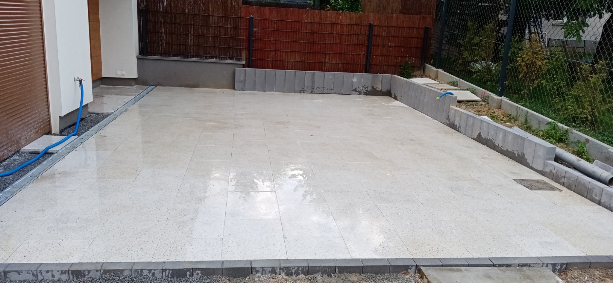 Wyłożony jasnymi płytami taras z odpływem liniowym przy ścianie budynku, obrzeża z kostki, w tle ogrodzenie z brązowej plecionki i zielenią.