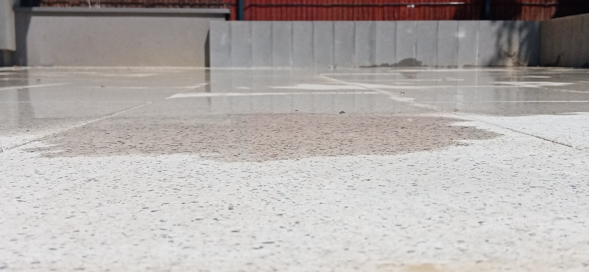 Powierzchnia z jasnoszarej kostki brukowej, częściowo mokra, odbijająca niebo, z widocznymi fugami i zarysem budynku w tle.