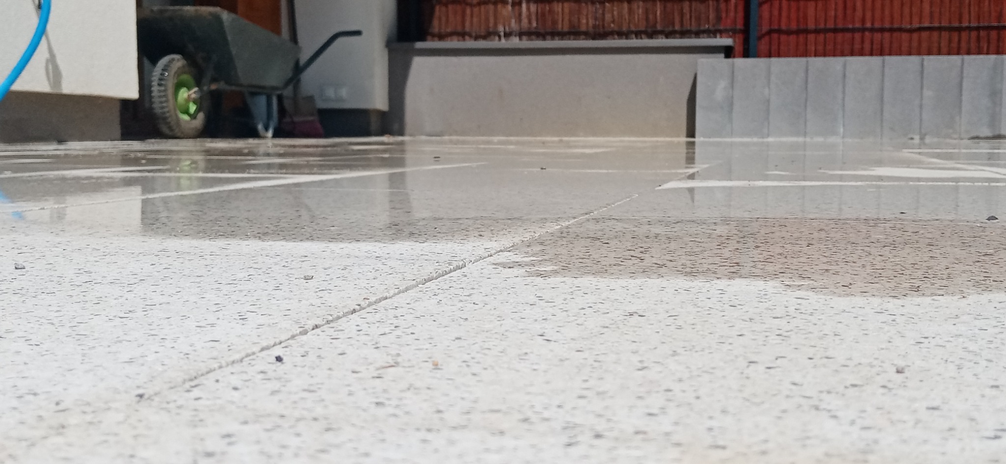 Świeżo ułożona, mokra nawierzchnia z jasnych płyt betonowych z widocznymi fugami i wózkiem budowlanym w tle.