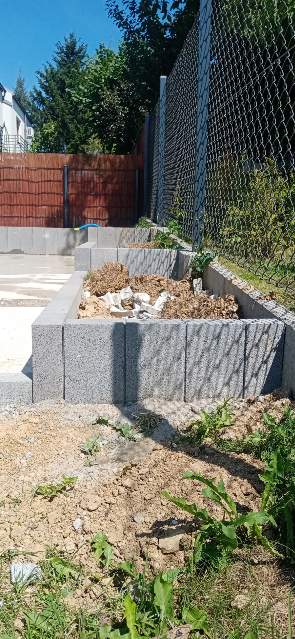 Wznoszenie murku oporowego z szarej kostki betonowej przy ogrodzeniu z siatki, widoczne pozostałości ziemi i gruzu wewnątrz konstrukcji, zarośnięty teren wokół.