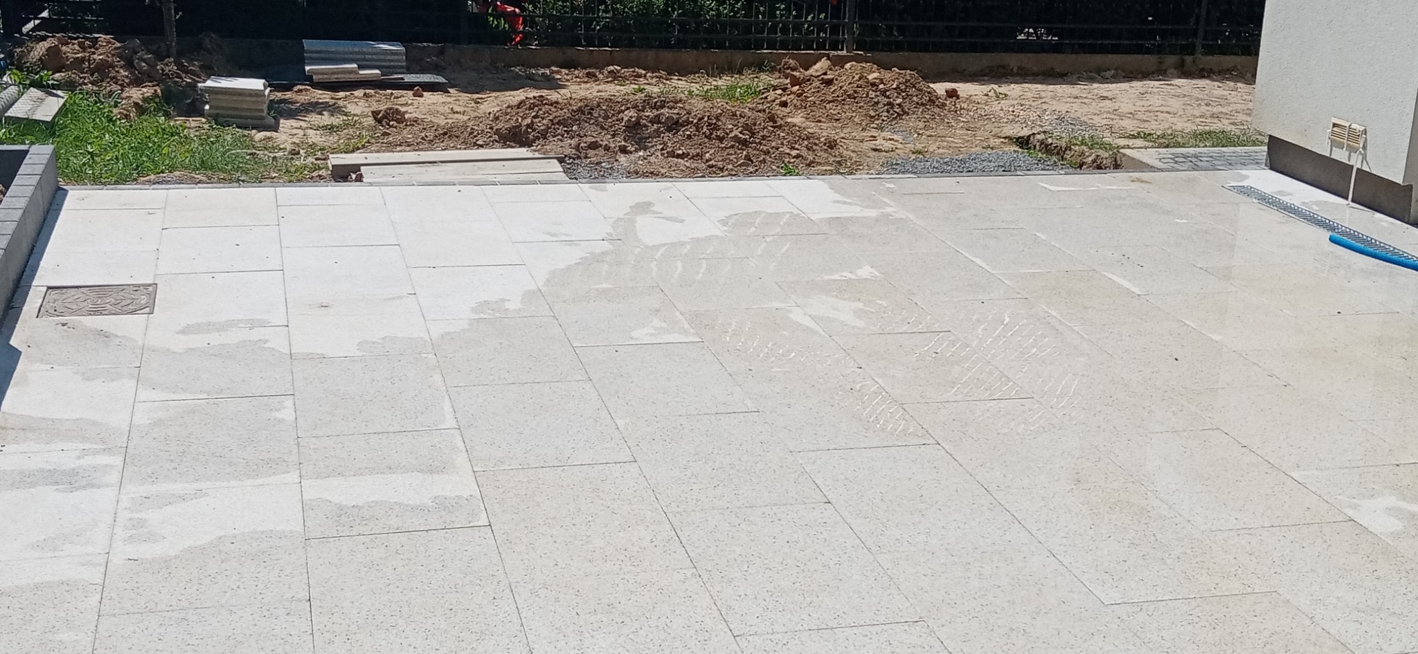 Jasne, prostokątne płyty chodnikowe ułożone na zewnątrz budynku, z widocznymi śladami wilgoci, studzienką kanalizacyjną i fragmentem odwodnienia liniowego przy ścianie budynku; w tle widoczny stos...