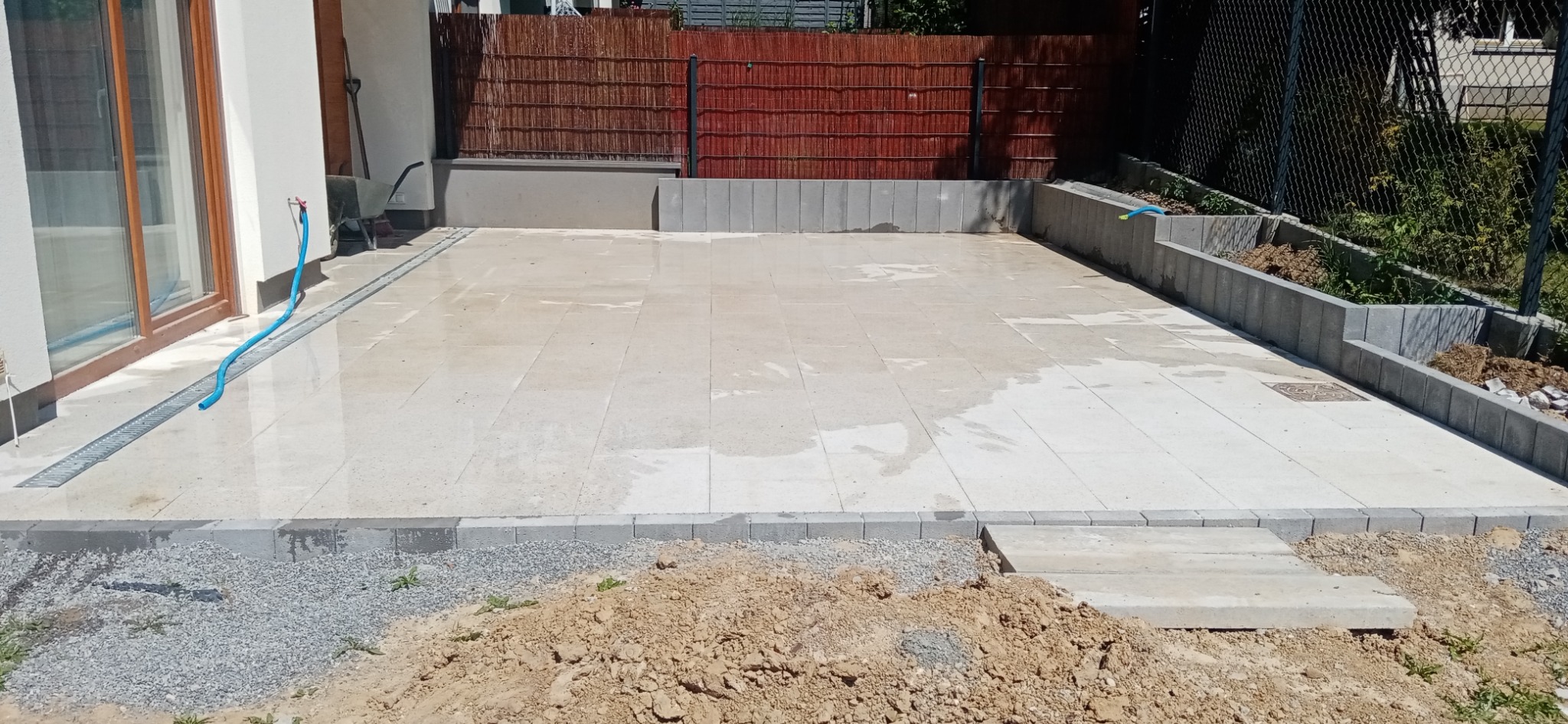 Ułożona jasna kostka brukowa na posesji, z widocznym odwodnieniem liniowym przy budynku, obrzeżami z kostki i schodkami wejściowymi, na tle ogrodzenia z siatki i roślinności.