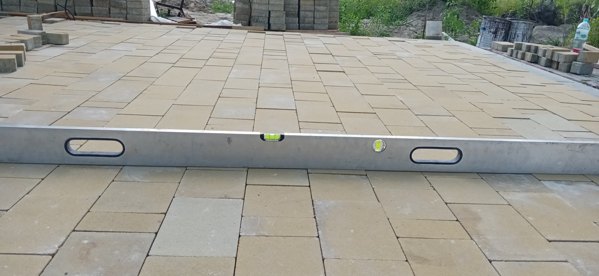 Poziomnica spoczywa na częściowo ułożonej, beżowej kostce brukowej; w tle sterta kostki i zieleń.