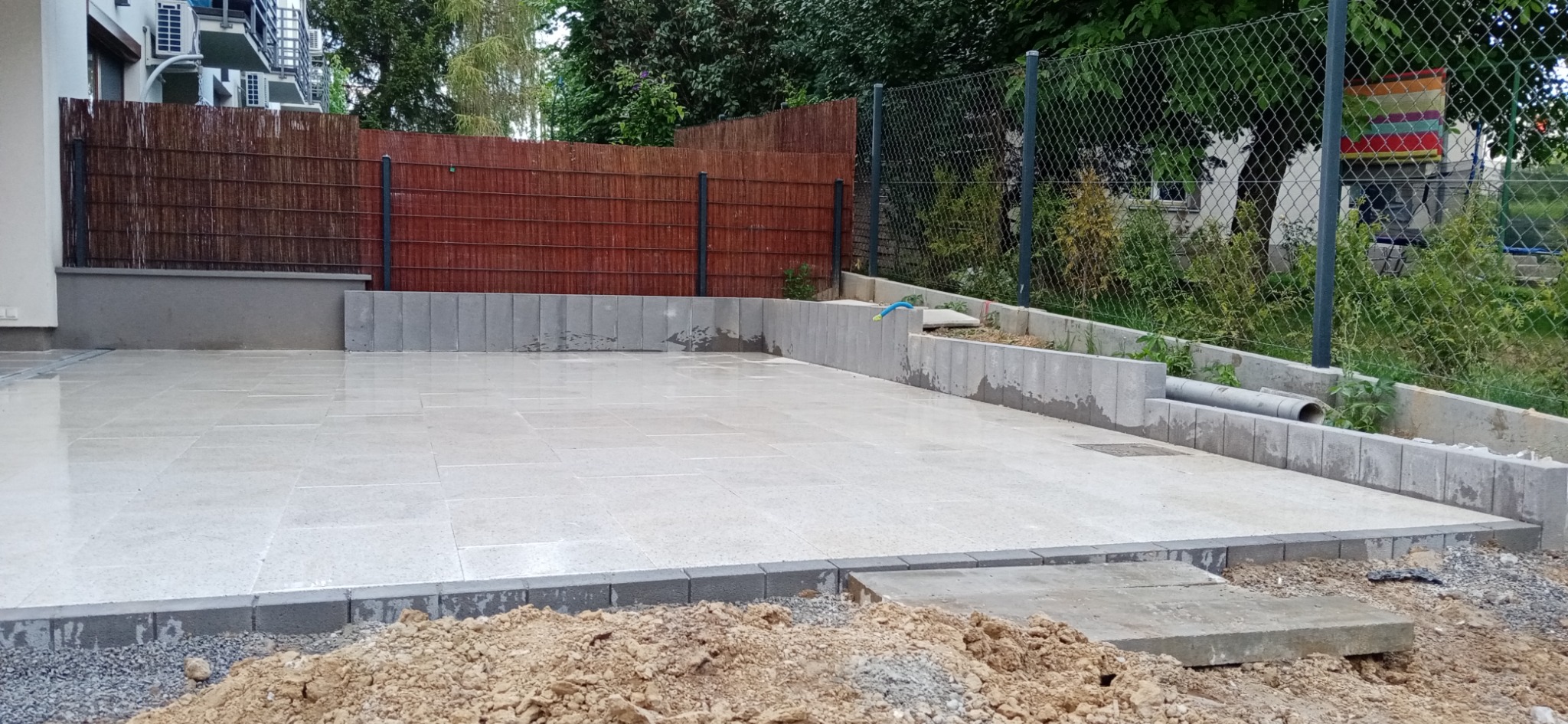 Wyłożony jasnymi płytami plac z obrzeżami z szarej kostki, widoczny fragment ziemi z przodu, w tle ogrodzenie z siatki i drewniana osłona, mokra powierzchnia po deszczu.