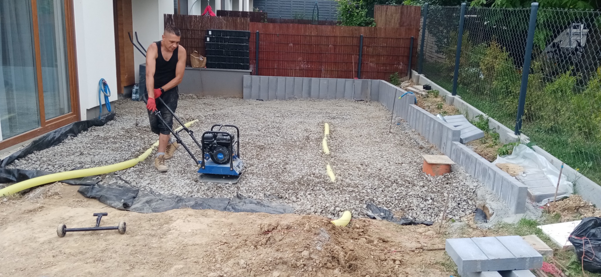 Pracownik ubija zagęszczarką wibracyjną podłoże z kamieni pod przyszłą nawierzchnię, widoczne krawężniki i obrzeża, przygotowanie terenu przed domem.
