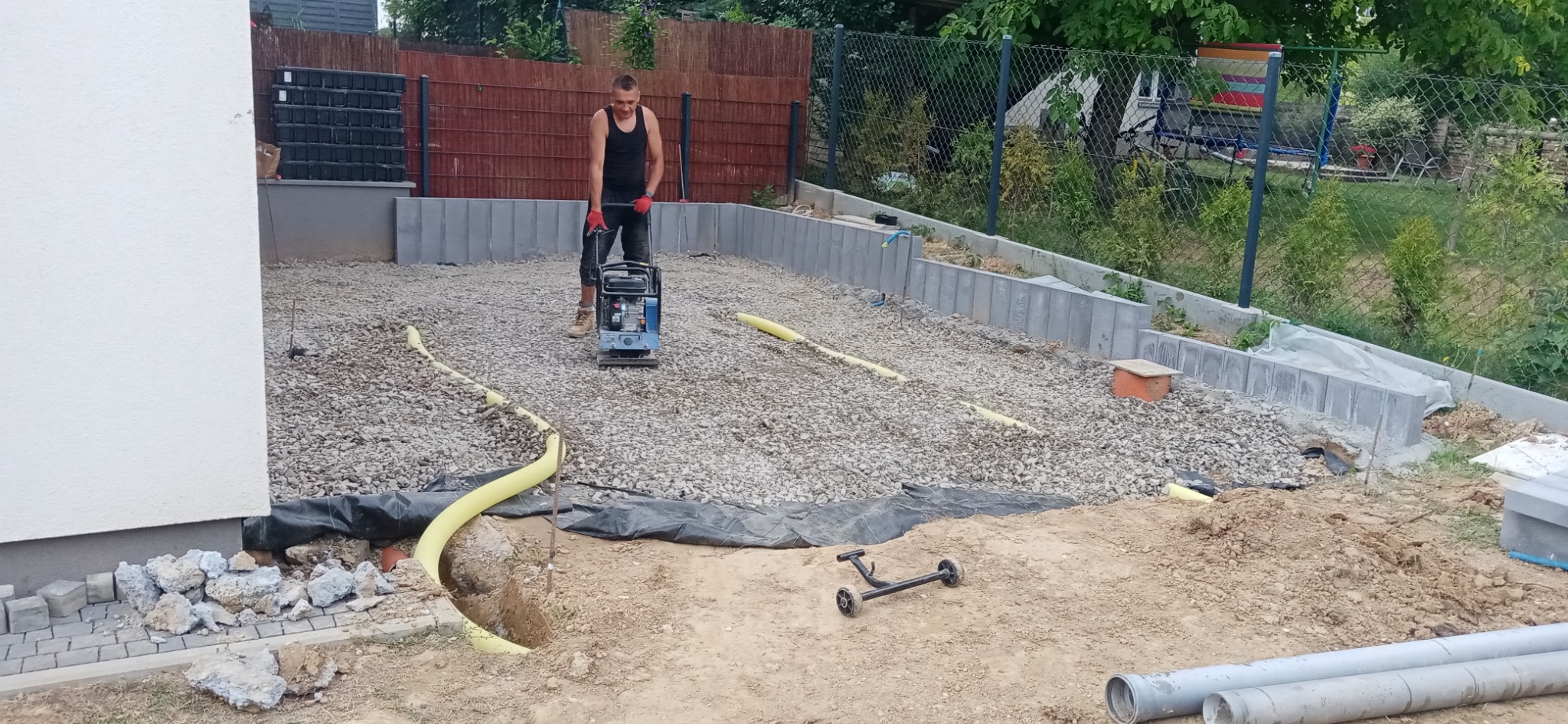 Prace ziemne: mężczyzna w roboczych butach i czerwonych rękawicach zagęszcza podłoże z kamieni za pomocą zagęszczarki wibracyjnej, widoczne rury i krawężniki.