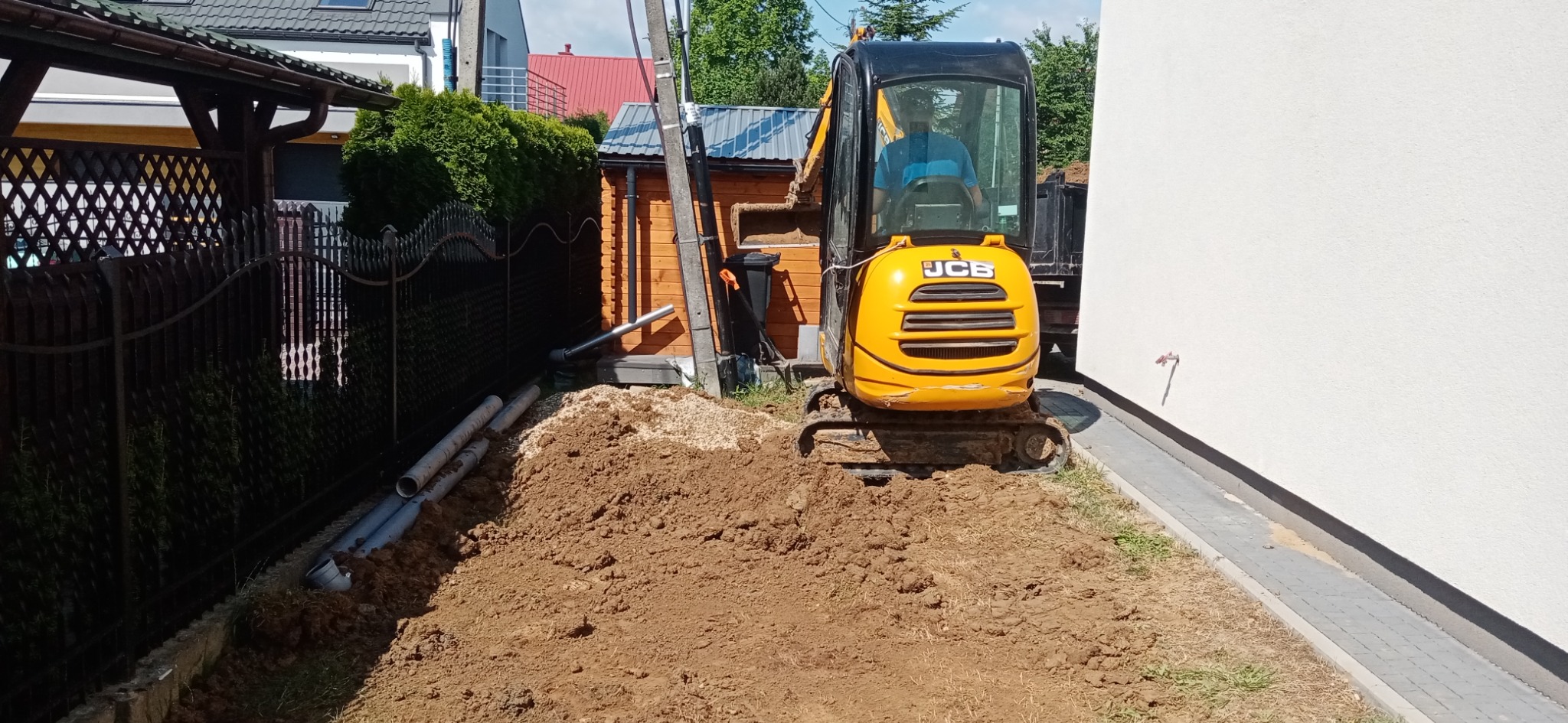 Żółta mini koparka JCB na placu budowy, obok czarnego ogrodzenia i rur kanalizacyjnych, na tle białej ściany budynku i drewnianej szopy.