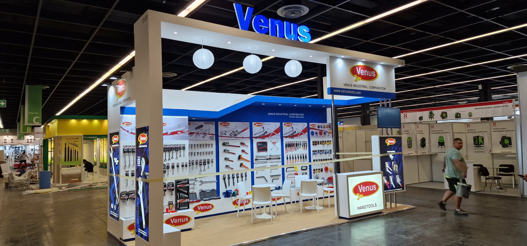 Stoisko targowe firmy Venus Industrial Corporation z ekspozycją narzędzi ręcznych, oświetlone kulistymi lampami, z widocznymi katalogami i narzędziami na ścianach oraz trzema białymi krzesłami...